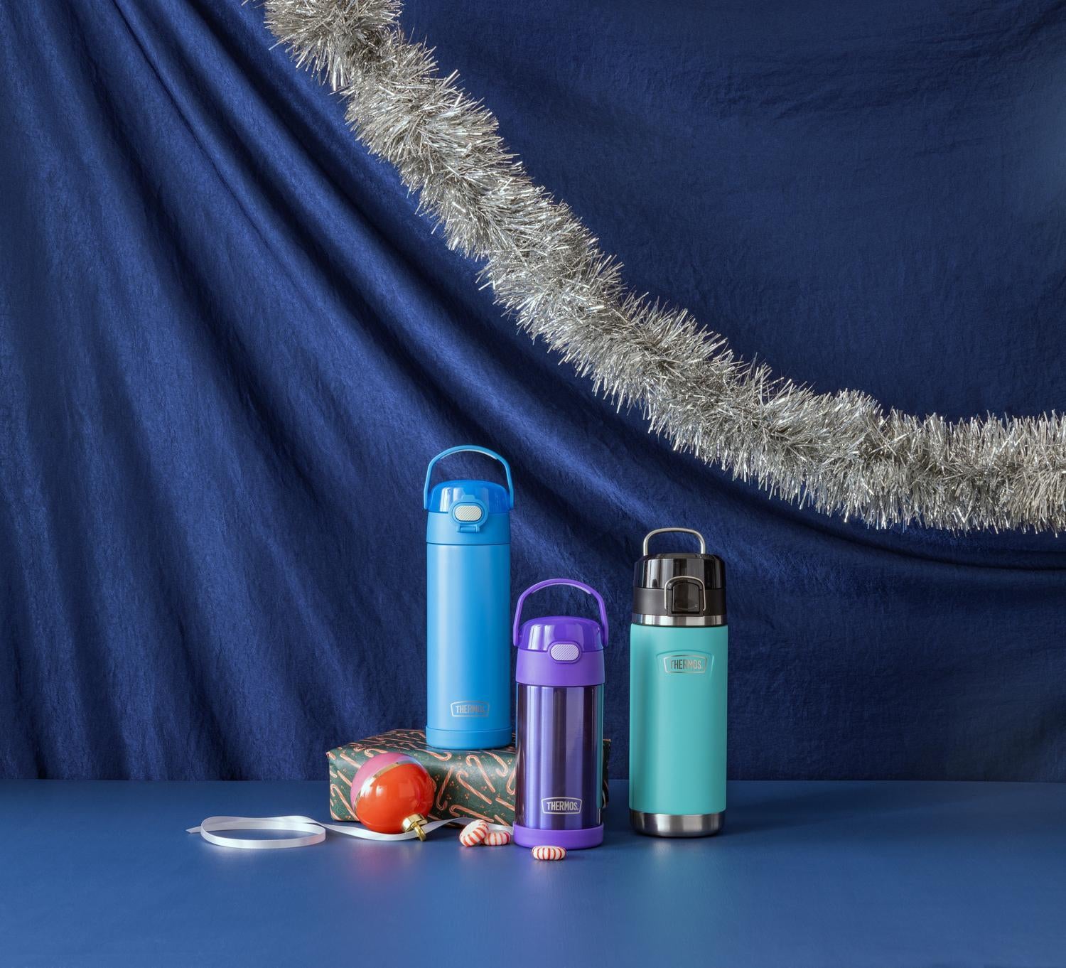 Three insulated bottles in blue, purple, and green on a blue surface with a festive tinsel border.