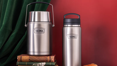 Two stainless steel thermos bottles on a red surface with a green curtain in the background