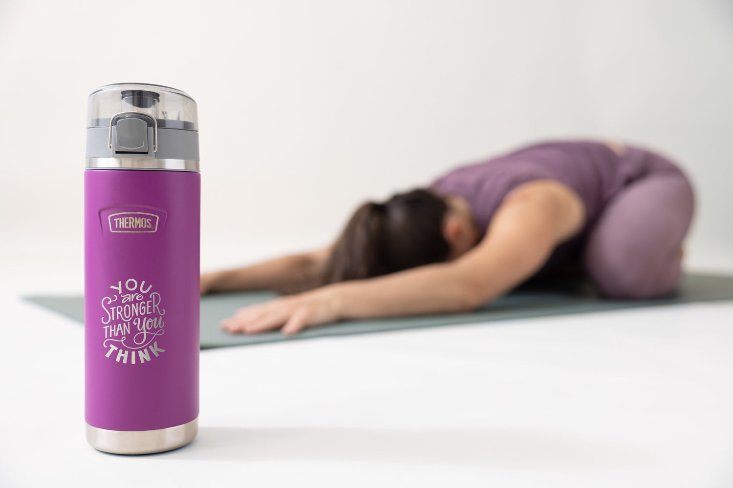 Purple thermos stainless steel water bottle with motivational quote on a white surface, with a blurred background of a person exercising.