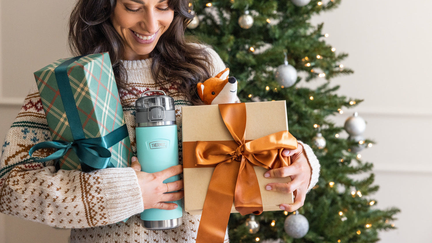 Woman holding a gift, thermos, and wrapped present in front of a Christmas tree.