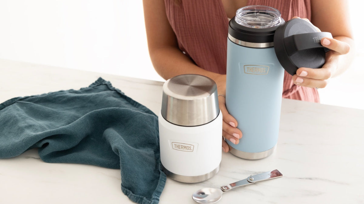 Person holding a blue thermos with a white thermos and spoon on a light surface.