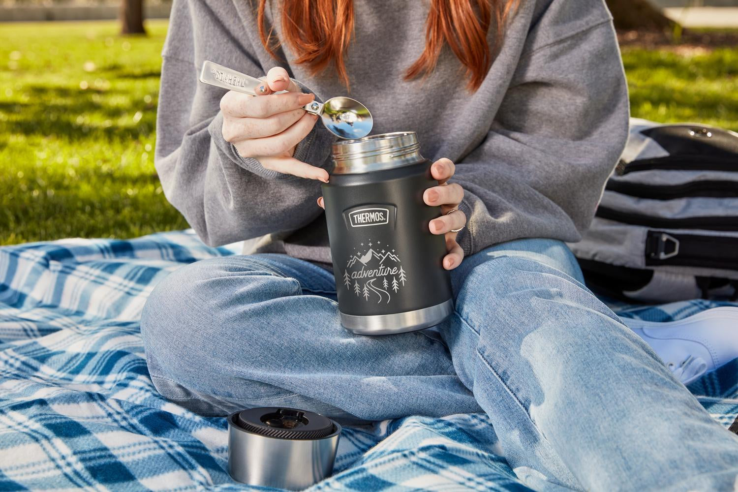 Person sitting on a blanket outdoors, holding and eating out of a black thermos with a forest design engraved on the front with the word "Adventure".