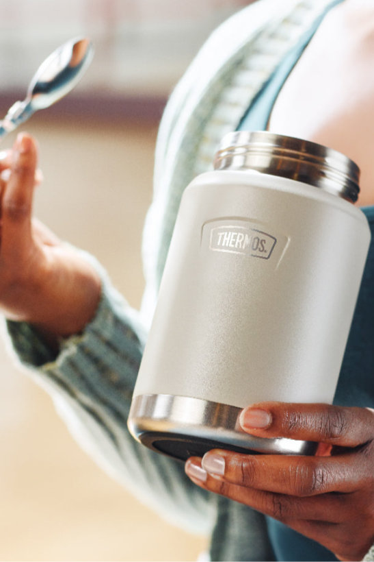 Person holding a white thermos food jar with a spoon, wearing a striped shirt.