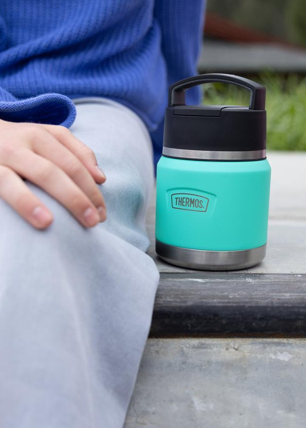 Thermos Turquoise thermos with black lid on a wooden surface, person wearing blue sweater and light pants in the background