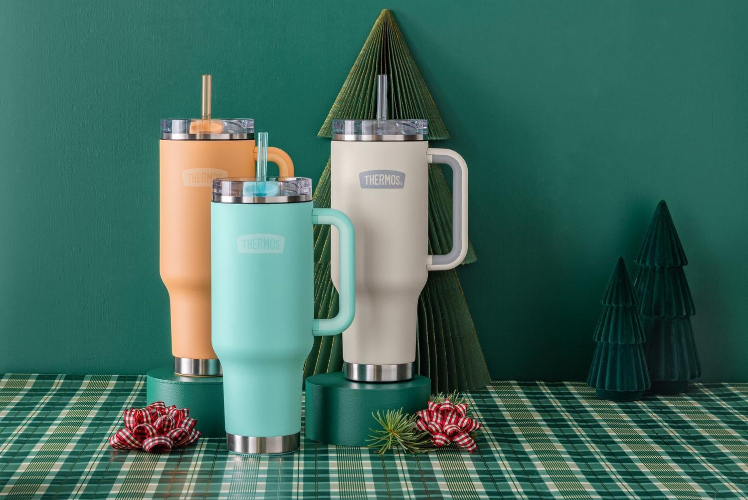 Set of colorful Thermos insulated mugs with straws on a green plaid tablecloth with decorative trees in the background.