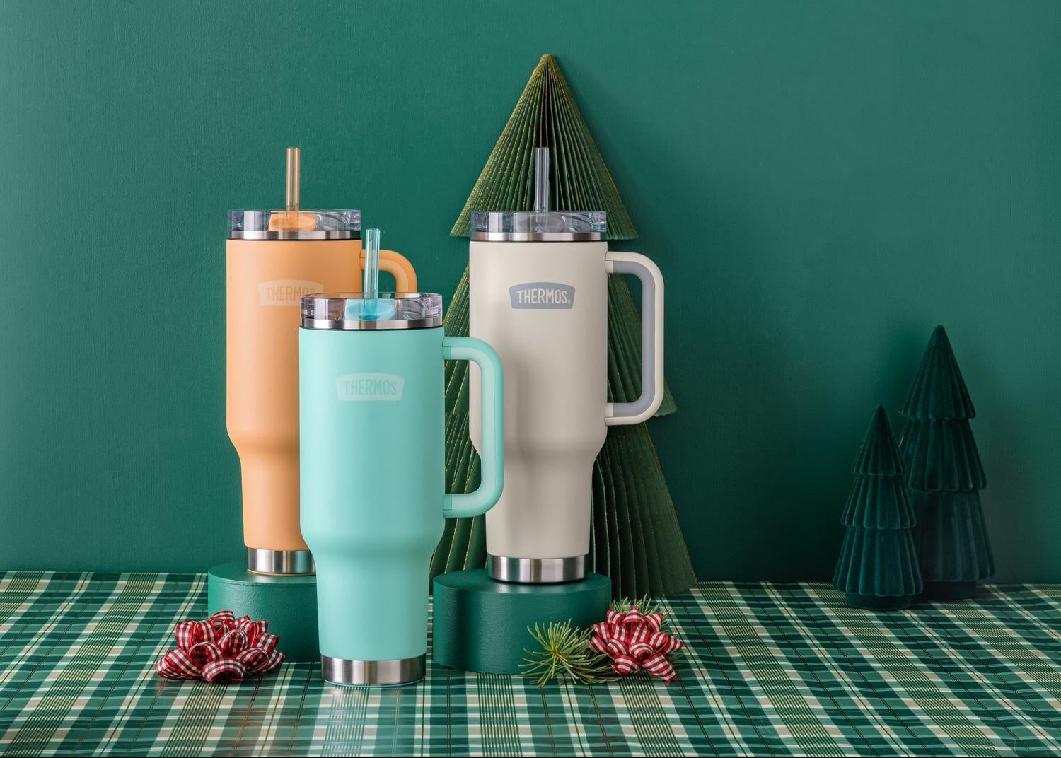 Set of colorful Thermos insulated mugs on a green plaid tablecloth with decorative trees in the background.